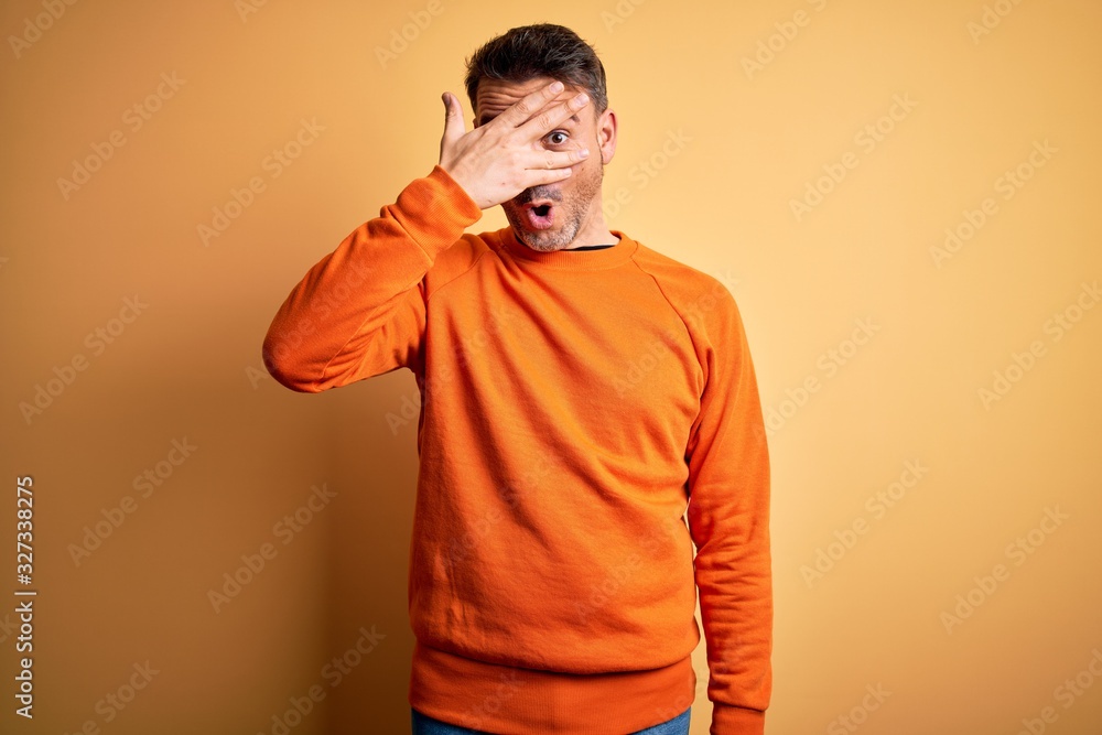 Young handsome man wearing orange casual sweater standing over isolated yellow background peeking in shock covering face and eyes with hand, looking through fingers with embarrassed expression. Young handsome man wearing orange casual sweater standing over isolated yellow background peeking in shock covering face and eyes with hand, looking through fingers with embarrassed expression.