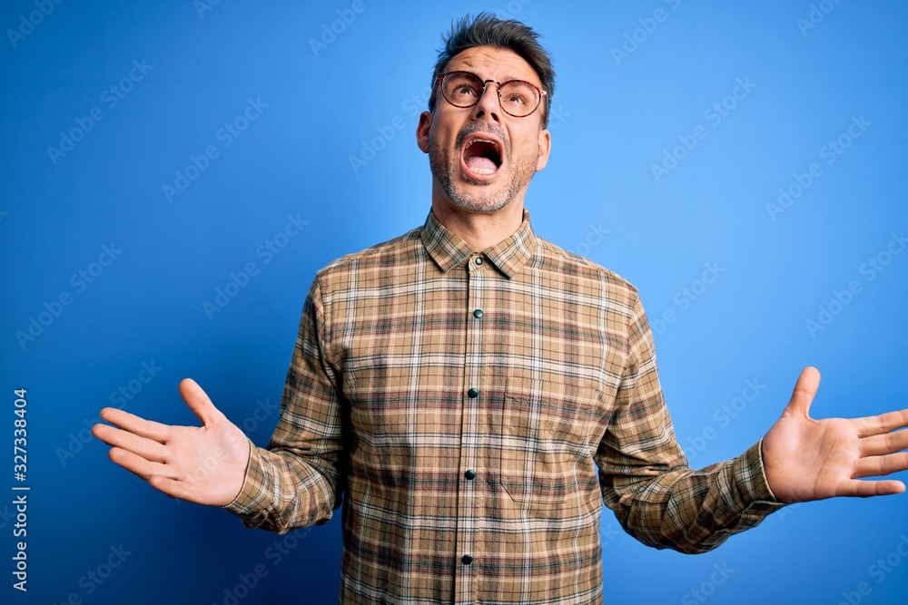 Fototapeta premium Young handsome man wearing casual shirt and glasses standing over blue background crazy and mad shouting and yelling with aggressive expression and arms raised. Frustration concept.