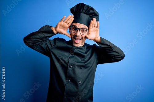 Young handsome chef man wearing cooker uniform and hat over isolated blue background Smiling cheerful playing peek a boo with hands showing face. Surprised and exited