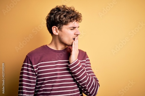 Young blond handsome man with curly hair wearing casual striped sweater bored yawning tired covering mouth with hand. Restless and sleepiness. © Krakenimages.com