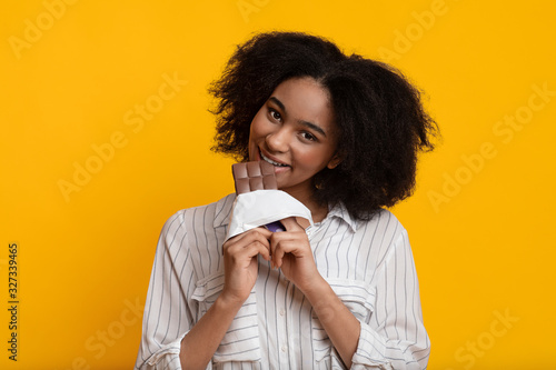 Happy afro woman eating chocolate bar, biting piece, smiling to camera