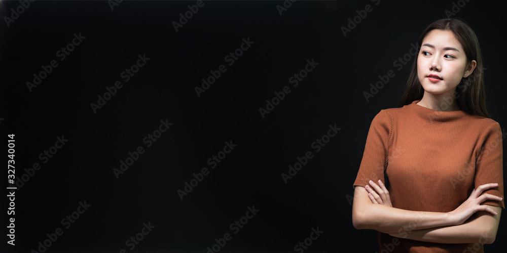 Portrait of young Asian woman wearing brown casual shirt cross her arms and rolling her eyes to her right hand side on the wide black screen and copy space. Portrait of young Asian woman wearing brown casual shirt cross her arms and rolling her eyes to her right hand side on the wide black screen and copy space.