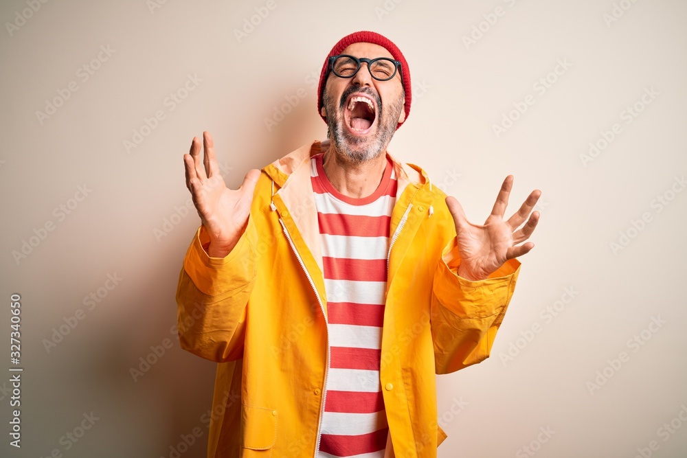 Middle age hoary man wearing glasses and rain coat standing over isolated white background crazy and mad shouting and yelling with aggressive expression and arms raised. Frustration concept.