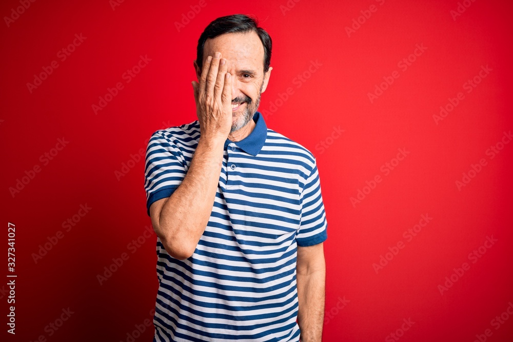 Middle age hoary man wearing casual striped polo standing over isolated red background covering one eye with hand, confident smile on face and surprise emotion. Middle age hoary man wearing casual striped polo standing over isolated red background covering one eye with hand, confident smile on face and surprise emotion.