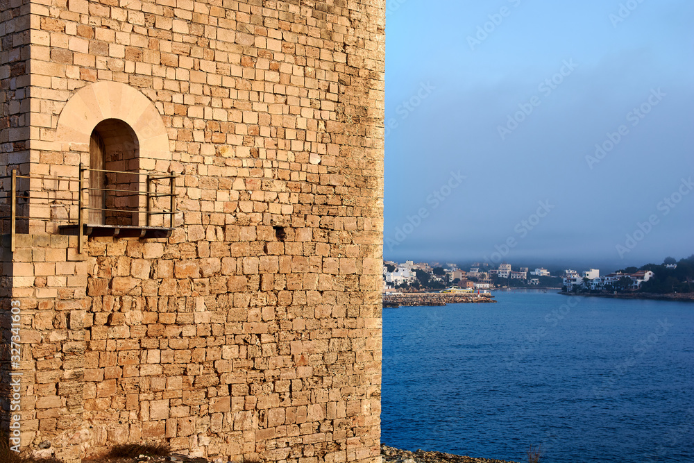 Mediterranean landscape on a sunny, foggy morning. Medieval tower in the foreground and small fishing village in the background. Porto Petro, Mallorca. Balearic Islands. Spain.