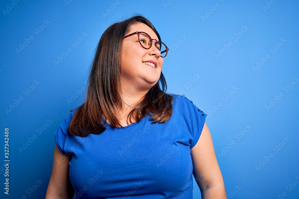 Beautiful brunette plus size woman wearing casual t-shirt over isolated blue background looking away to side with smile on face, natural expression. Laughing confident. Beautiful brunette plus size woman wearing casual t-shirt over isolated blue background looking away to side with smile on face, natural expression. Laughing confident.