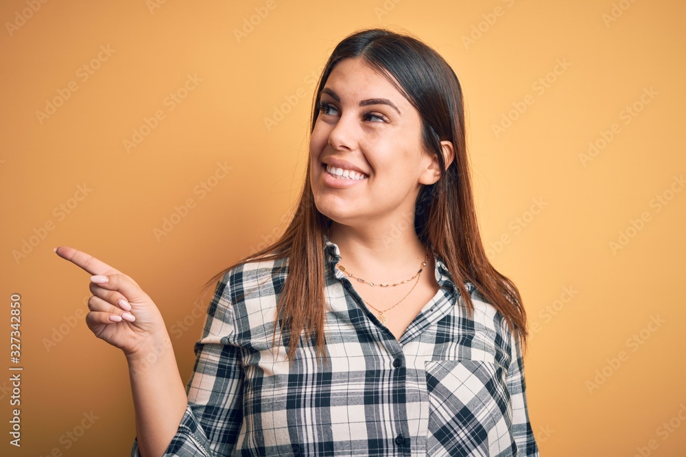 Young beautiful woman wearing casual shirt standing over isolated orange background with a big smile on face, pointing with hand and finger to the side looking at the camera. Young beautiful woman wearing casual shirt standing over isolated orange background with a big smile on face, pointing with hand and finger to the side looking at the camera.