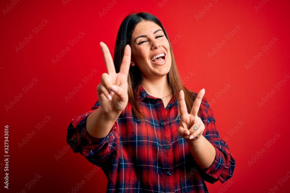Young beautiful woman wearing casual shirt over red background smiling looking to the camera showing fingers doing victory sign. Number two. Young beautiful woman wearing casual shirt over red background smiling looking to the camera showing fingers doing victory sign. Number two.
