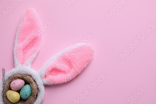 Headband with Easter bunny ears and dyed eggs in nest on pink background, flat lay. Space for text