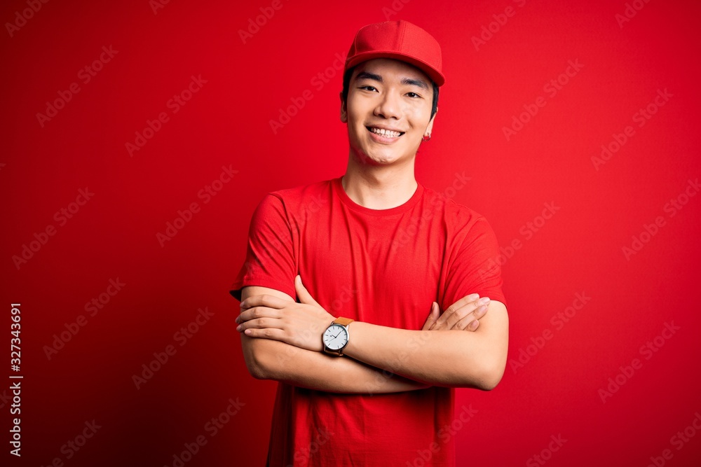 Young handsome chinese delivery man wearing cap standing over isolated red background happy face smiling with crossed arms looking at the camera. Positive person. Young handsome chinese delivery man wearing cap standing over isolated red background happy face smiling with crossed arms looking at the camera. Positive person.