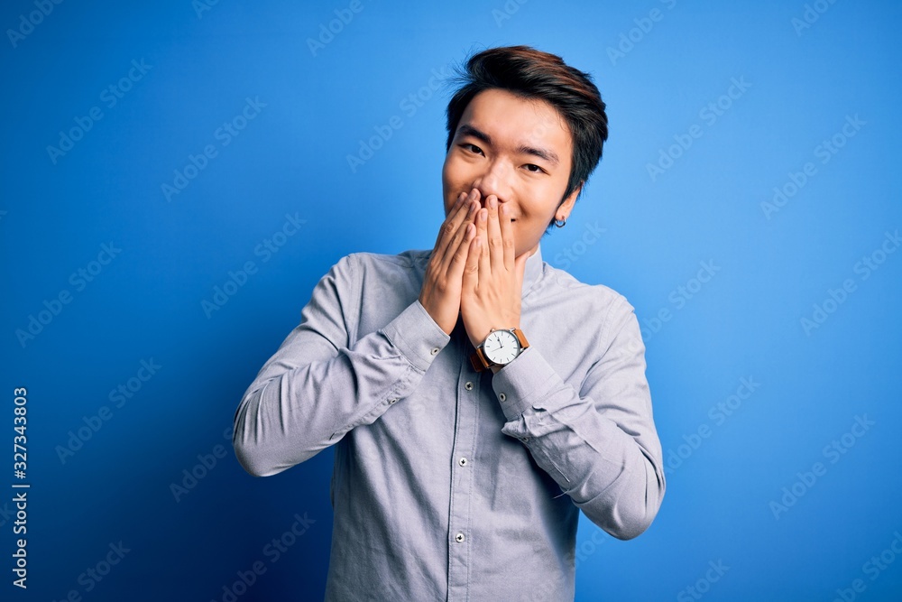 Young handsome chinese man wearing casual shirt standing over isolated blue background laughing and embarrassed giggle covering mouth with hands, gossip and scandal concept Young handsome chinese man wearing casual shirt standing over isolated blue background laughing and embarrassed giggle covering mouth with hands, gossip and scandal concept