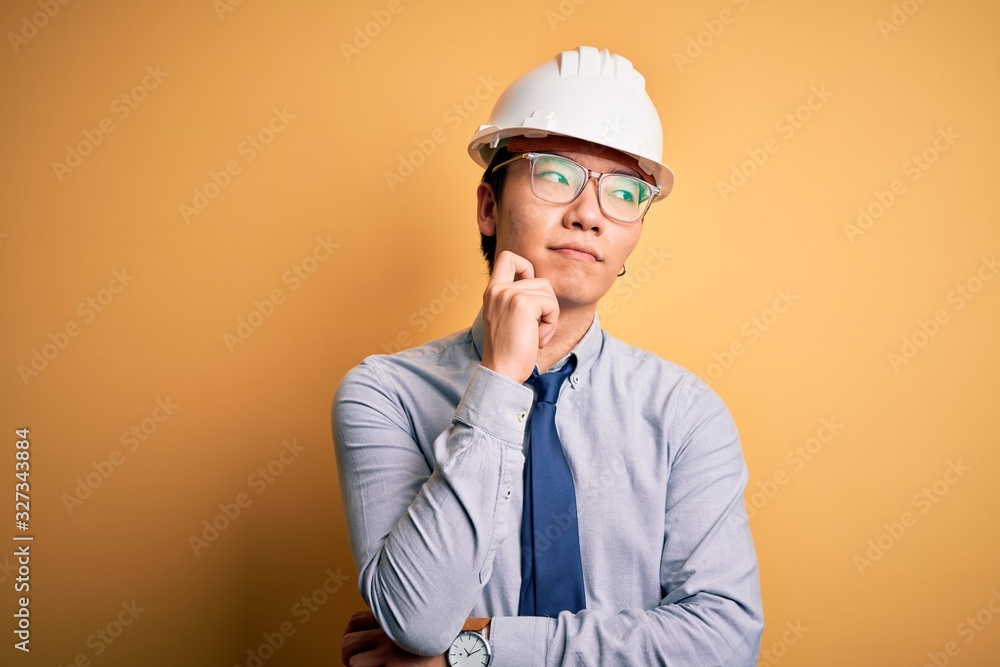 Young handsome chinese architect man wearing safety helmet and tie over yellow background with hand on chin thinking about question, pensive expression. Smiling with thoughtful face. Doubt concept.