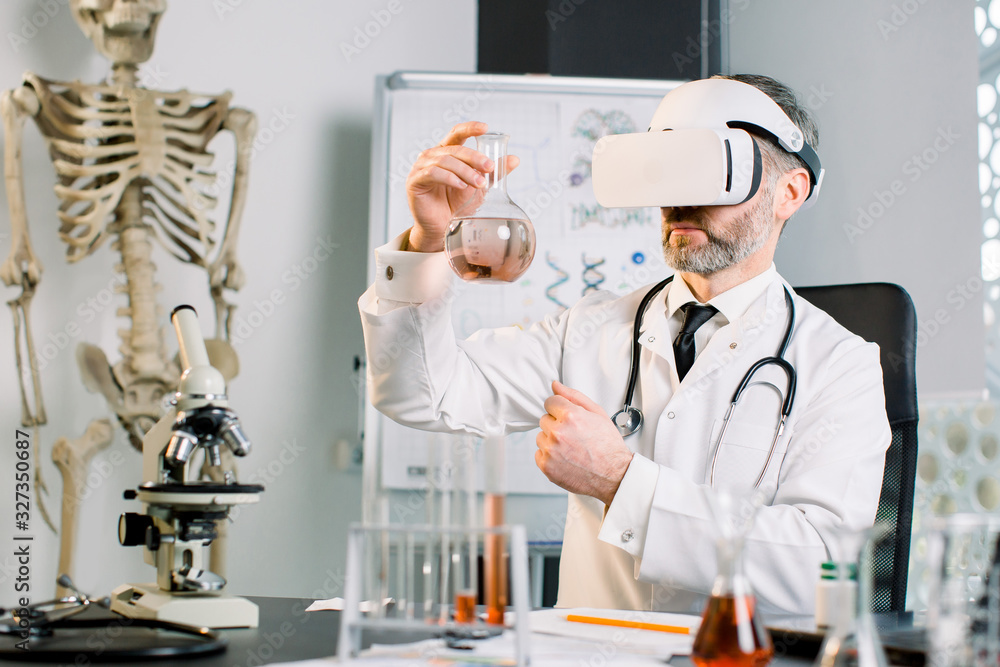 Man scientist investigator checking new liquid in glass flask, using vr glasses, working at the table in modern pharmaceutical lab. Science, chemistry, biology, medicine and people concept Man scientist investigator checking new liquid in glass flask, using vr glasses, working at the table in modern pharmaceutical lab. Science, chemistry, biology, medicine and people concept
