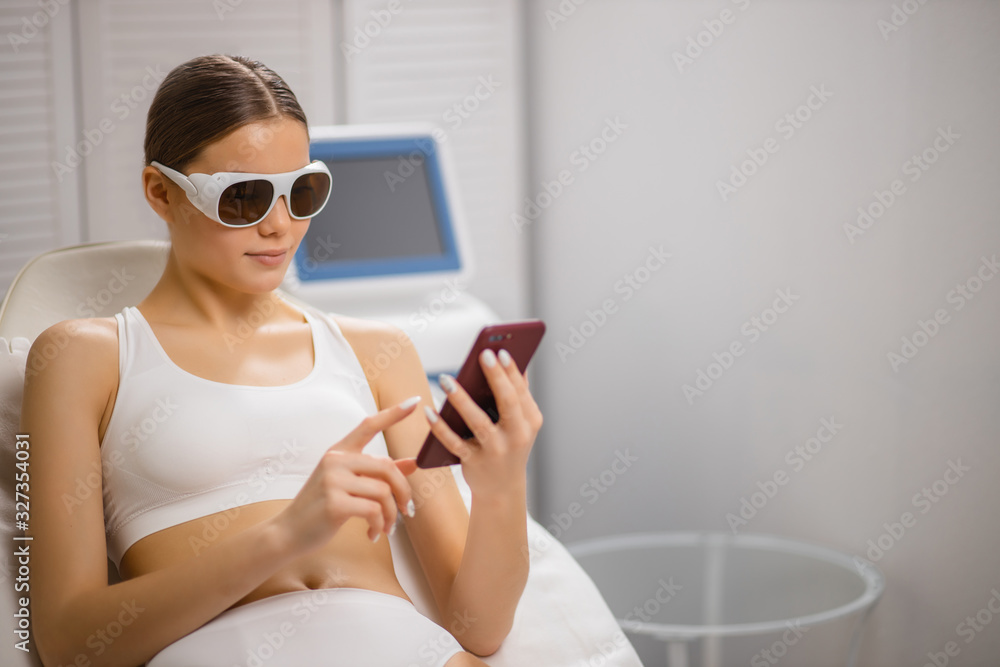 caucasian woman at laser epilation procedure, use smartphone while getting hair removal procedure in beauty salon, wearing protective eyeglasses. hairless, hair removal, epilation, depilation concept