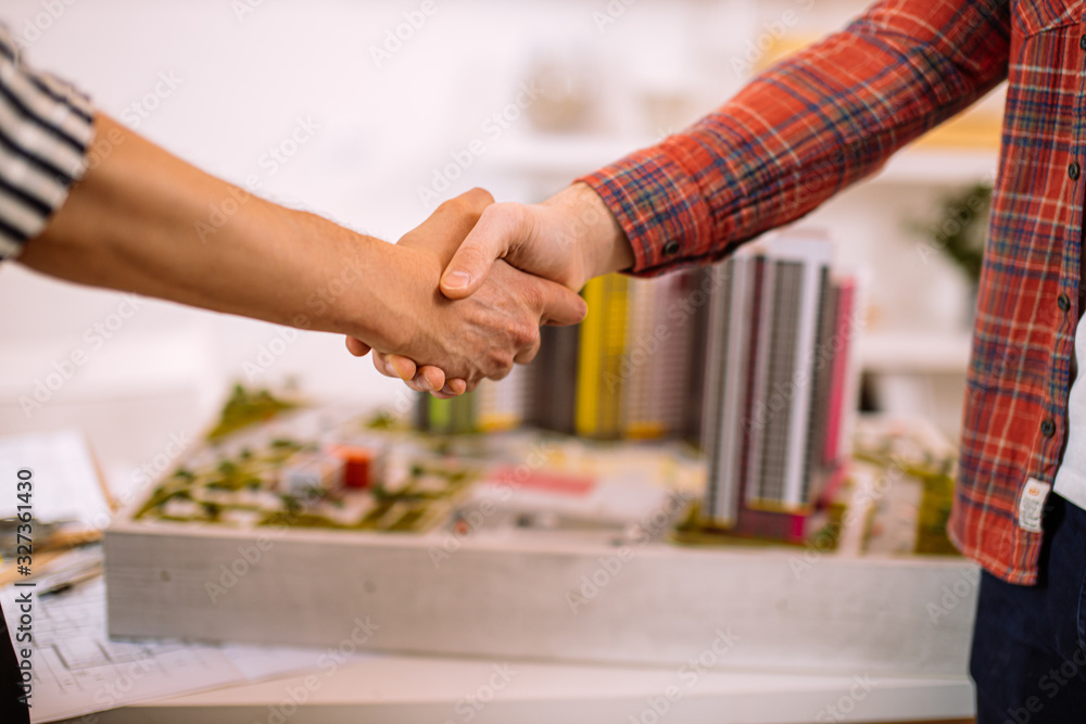 two young successful architects shaking hands in office above high-rise building model, they signed a cooperation contract to build new residential house two young successful architects shaking hands in office above high-rise building model, they signed a cooperation contract to build new residential house