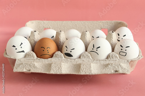 Group of white organic chicken eggs with angry faces and one brown chicken egg with crying face in carton box made from recycled paper. Red background. Theme of racism and intolerance in society.