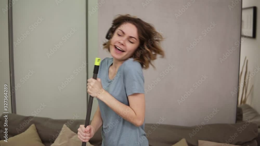 People, housework and housekeeping concept - happy woman in headphones with mop cleaning floor and dancing at home. Using swab pipe as a microphone. Grey couch on the background People, housework and housekeeping concept - happy woman in headphones with mop cleaning floor and dancing at home. Using swab pipe as a microphone. Grey couch on the background