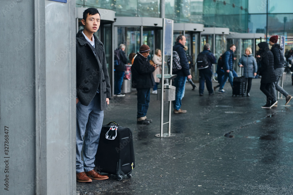 Young stylish Asian businessman waiting taxi while standing with suitcase near airport