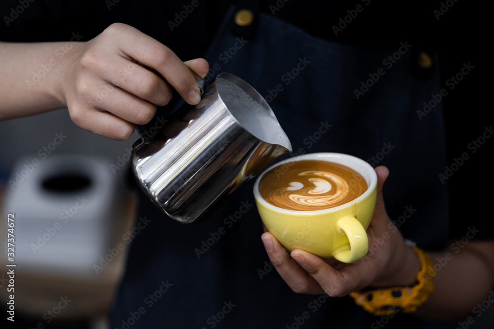 Close-up of barista hand holding and pouring hot milk for prepare latte art on a yellow cup of cappuccino coffee. Close-up of barista hand holding and pouring hot milk for prepare latte art on a yellow cup of cappuccino coffee.