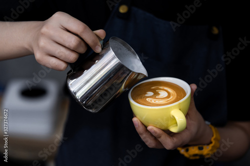 Close-up of barista hand holding and pouring hot milk for prepare latte art on a yellow cup of cappuccino coffee.