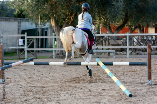 Back side of Girl riding a white horse jumping a guided obstacle during equestrian school training © daniele russo
