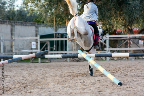 Back side of Girl riding a white horse jumping a guided obstacle during equestrian school training