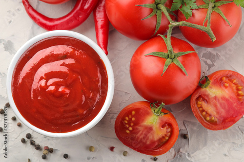 Red sauce or ketchup in a bowl and ingredients for cooking, peppers, spices, tomatoes on a light concrete background.