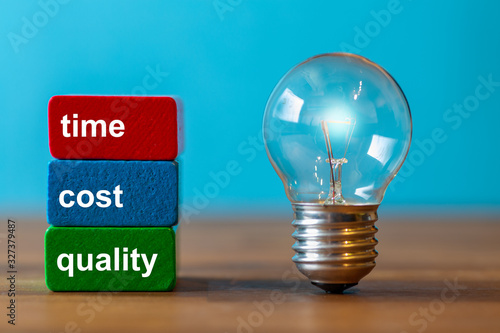 Time, cost quality stands on colorful blocks next to a vintage light bulb on a dark wooden plate with a blue background