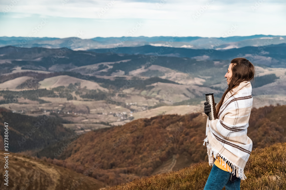 Scenery autumn sunny day. Woman standing covered in plaid with termo cup in hand watching the mountains peak, sky with clouds.