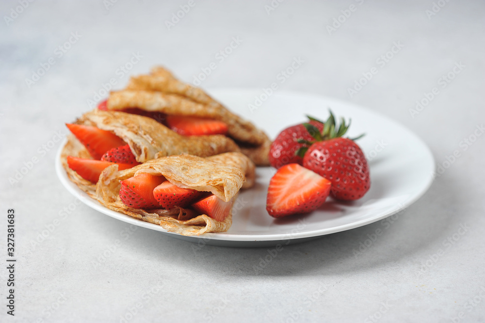 Thin pancakes with slices of strawberries. A few strawberries on a plate next to pancakes. Close-up. Light background. Thin pancakes with slices of strawberries. A few strawberries on a plate next to pancakes. Close-up. Light background.
