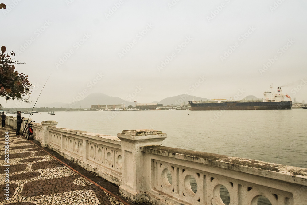 View of Santos city waterfront with the iconic white railing in the foreground and a ship navigating the Santos estuary in the background on a cloudy day. View of Santos city waterfront with the iconic white railing in the foreground and a ship navigating the Santos estuary in the background on a cloudy day.