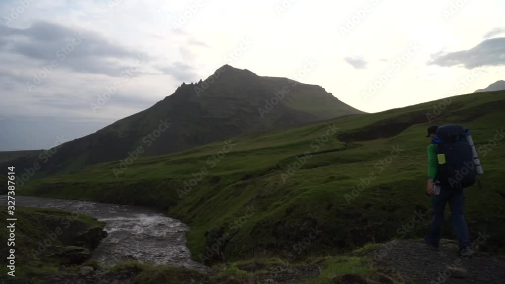 Young hiker come closer to river and green mountains on background on the Fimmvorduhals hiking trail in Iceland. Concept of freedom movement and freedom Young hiker come closer to river and green mountains on background on the Fimmvorduhals hiking trail in Iceland. Concept of freedom movement and freedom