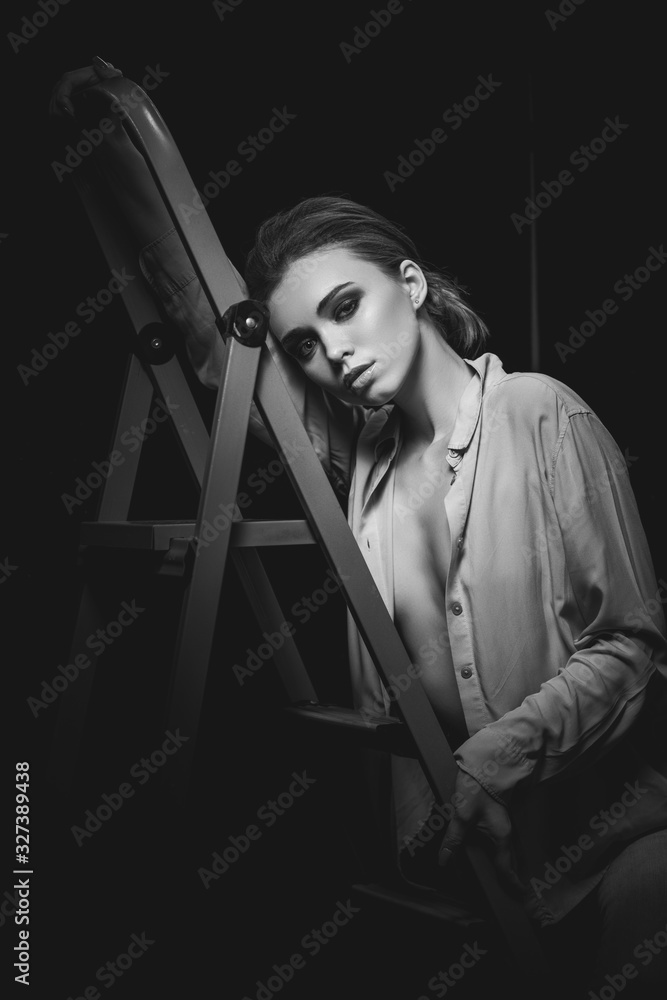 Obraz premium Portrait of stylish fashionable brunette woman model in gray shirt posing on ladder on black studio background.