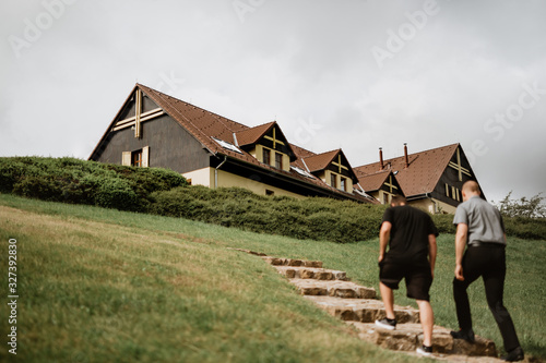 Grassy hill with big cottage on top. The cottage has wooden arcades and decorated roof gables. In the foreground, out of focus, two men are walking towards the cottage up the rocky stairs.