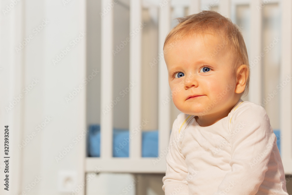 Close portrait of beautiful baby boy at toddler age with calm face expression sitting near crib