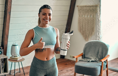 Athletic woman in sportswear holding a protein shake or a bottle of water and showing thumbs up gesture at home in the living room. © Valerii Apetroaiei