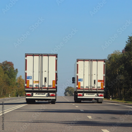 Two semi trucks with white van on suburban asphalt highway road at Sunny summer day against the blue sky and green forest on roadside, operative international cargo logistics spedition in Europe