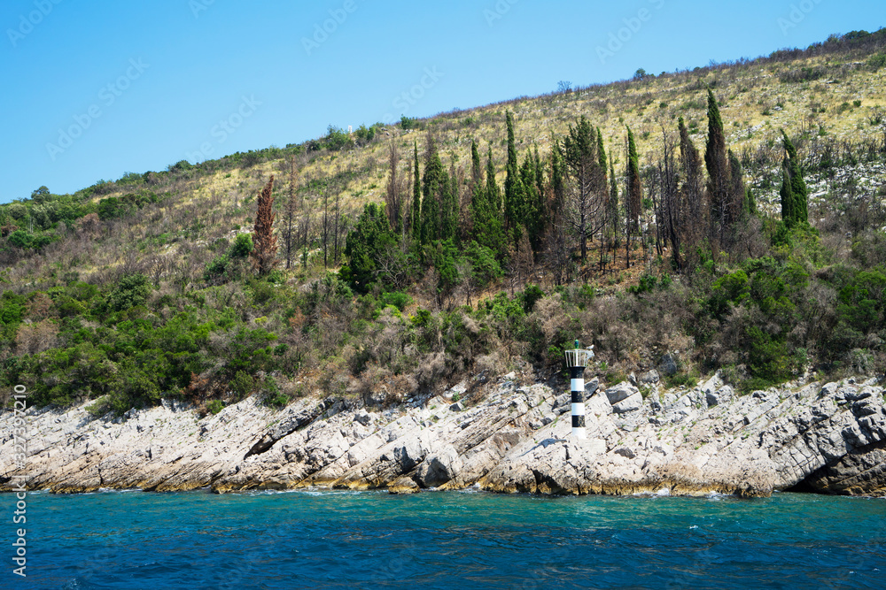 The rocky coast of the sea with a small black and white lighthouse and a forest with a cloudless sky on a sunny summer day. Kotor Bay. Montenegro.