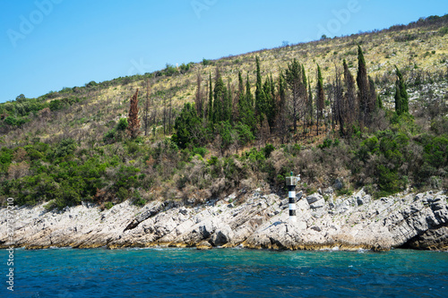 The rocky coast of the sea with a small black and white lighthouse and a forest with a cloudless sky on a sunny summer day. Kotor Bay. Montenegro.