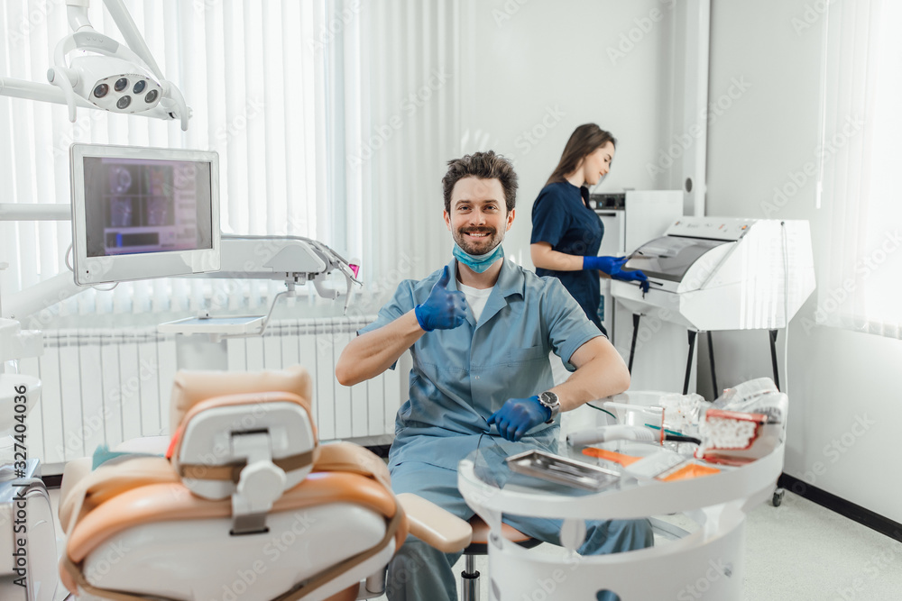 Young handsome and bearded dentist holding folder, while his assistant nurse preparing equipment. Ready for work. Young handsome and bearded dentist holding folder, while his assistant nurse preparing equipment. Ready for work.