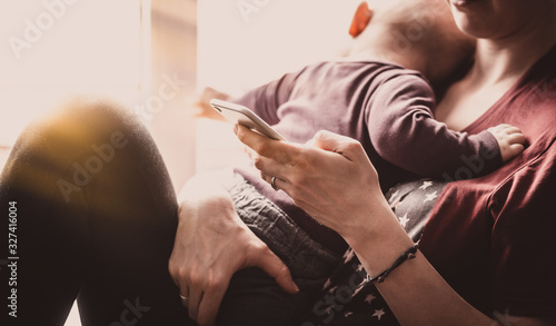 Female millennials holding a baby and a smartphone. Young woman looks after a young child and uses a smartphone. Phone is used in social media or for work via the Internet.