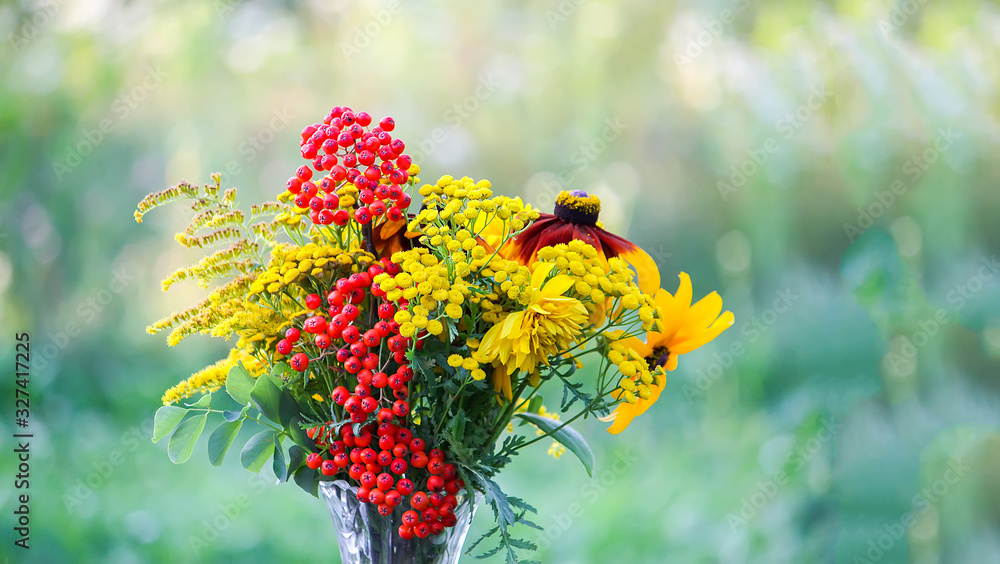 Handmade bouquet of wildflowers. Rowan berries on branch, goldenrog plant and tansy flowers. Floral composition in transparent vase on summer nature background outdoors. Handmade bouquet of wildflowers. Rowan berries on branch, goldenrog plant and tansy flowers. Floral composition in transparent vase on summer nature background outdoors.