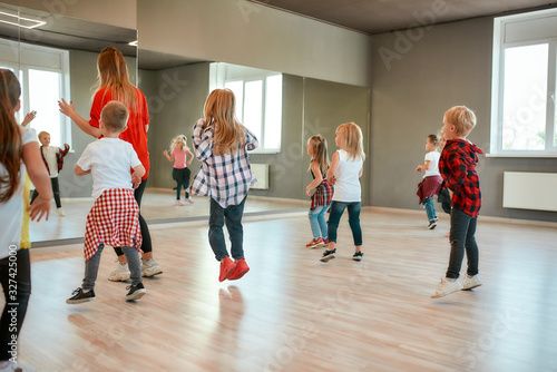 Fototapeta Naklejka Na Ścianę i Meble -  Choreography class. Group of active children dancing in front of the large mirror while having choreography class in the dance studio. Female dance teacher and kids