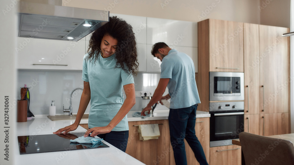 Obraz premium Clean Home. Two young professional cleaners in uniform working together in the kitchen. Happy afro american woman cleaning electric stove, young caucasian man wiping kitchen table