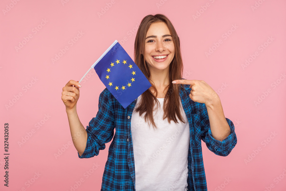 Portrait of positive friendly woman in checkered shirt smiling broadly and pointing at European Union flag, symbol of Europe, EU association, community. indoor studio shot isolated on pink background Portrait of positive friendly woman in checkered shirt smiling broadly and pointing at European Union flag, symbol of Europe, EU association, community. indoor studio shot isolated on pink background