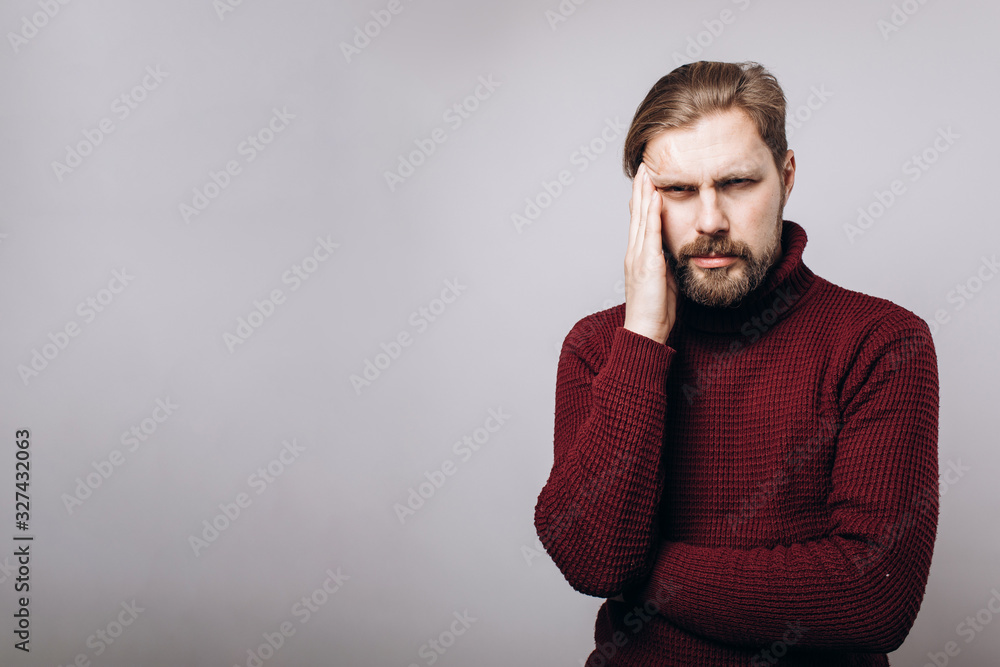 Middle-aged bearded man in claret-colored sweater suffering from headache while posing in studio over grey background. Mature male holding hand on head because of strong pain. Middle-aged bearded man in claret-colored sweater suffering from headache while posing in studio over grey background. Mature male holding hand on head because of strong pain.