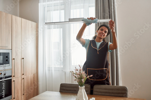 Perfectly clean. Young positive afro american house maid in apron wiping dust off a lamp while working in the modern kitchen. Cleaning services concept