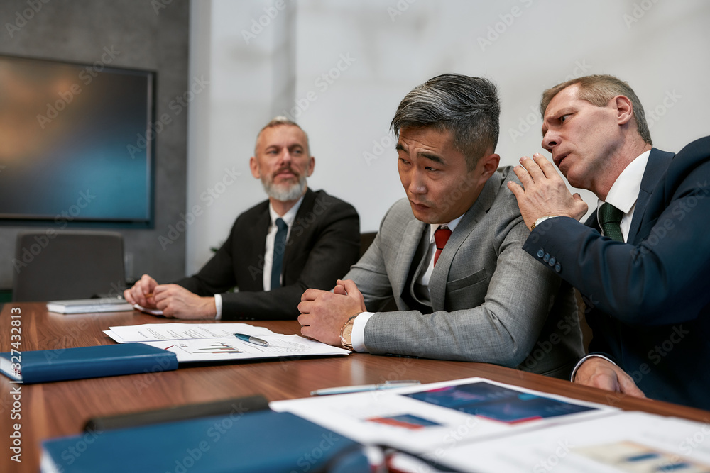Discussing contract conditions. Caucasian mature businessman whispering something to asian colleague in his ear while having a meeting in the modern office. Business people working together Discussing contract conditions. Caucasian mature businessman whispering something to asian colleague in his ear while having a meeting in the modern office. Business people working together