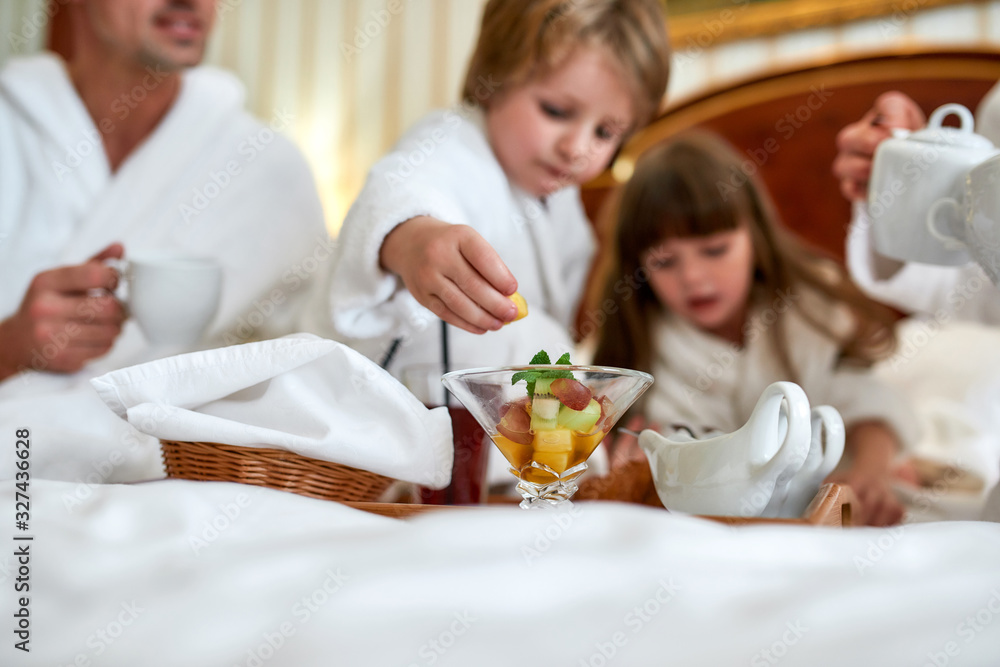 Eat good. Family in white bathrobes having breakfast in bed. Focus on fruit cup on the tray. Healthy food, resort, room service concept