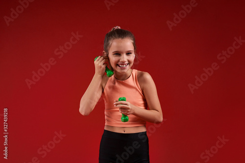 Having fun. A cute kid, girl is engaged in sport, she is smiling at camera while exercising with dumbbells. Isolated on red background. Fitness, training, active lifestyle concept. Horizontal shot